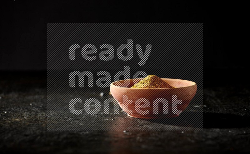 A wooden bowl full of turmeric powder with dried turmeric fingers on textured black flooring