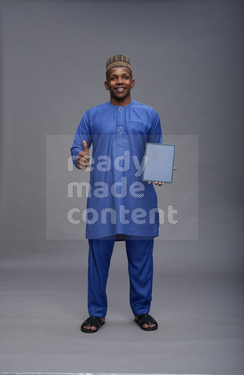 Man wearing Nigerian outfit standing showing tablet to camera on gray background