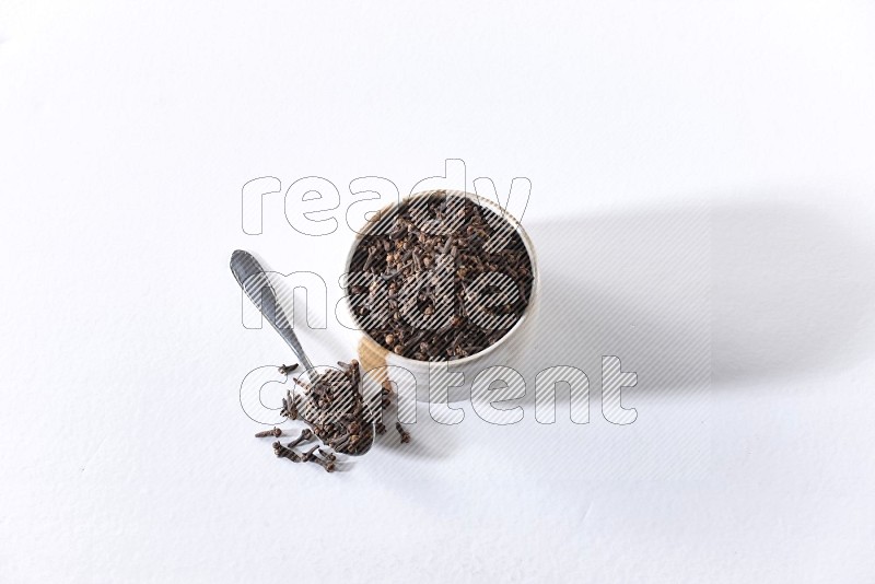 A beige ceramic bowl full of cloves and a metal spoon next to it on a white flooring