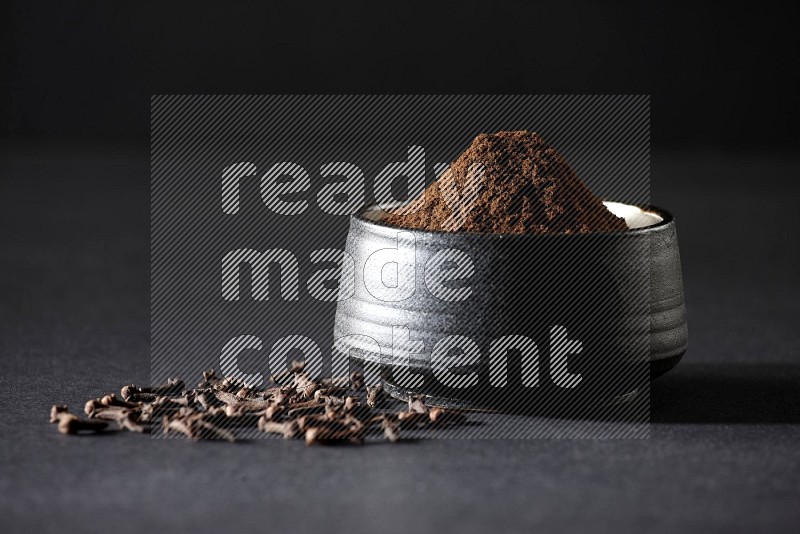 A black pottery bowl full of cloves powder and cloves whole grains on a black flooring