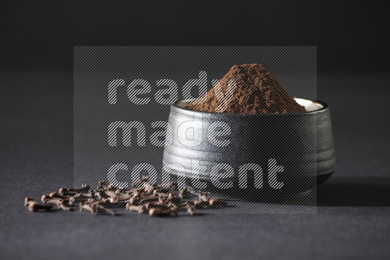 A black pottery bowl full of cloves powder and cloves whole grains on a black flooring