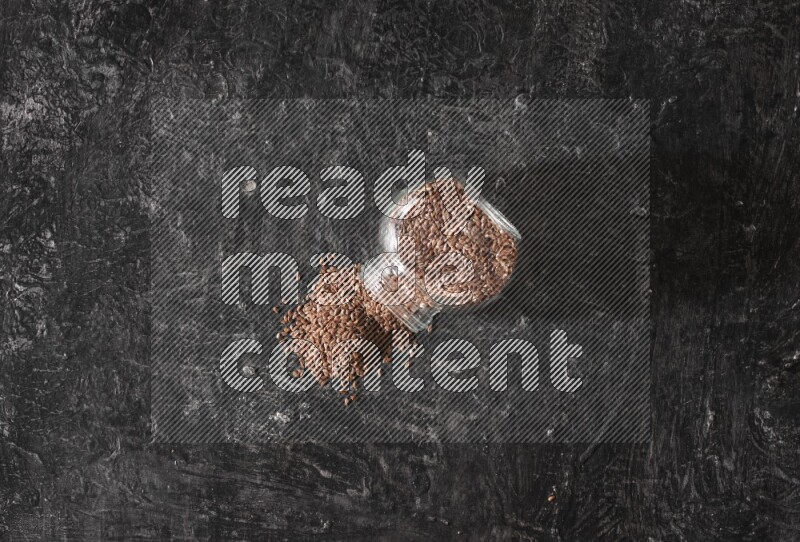 A glass spice jar full of flaxseeds flipped and seeds spread out on a textured black flooring