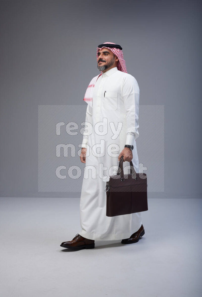 Saudi man Wearing Thob and red Shomag standing holding bag on Gray background
