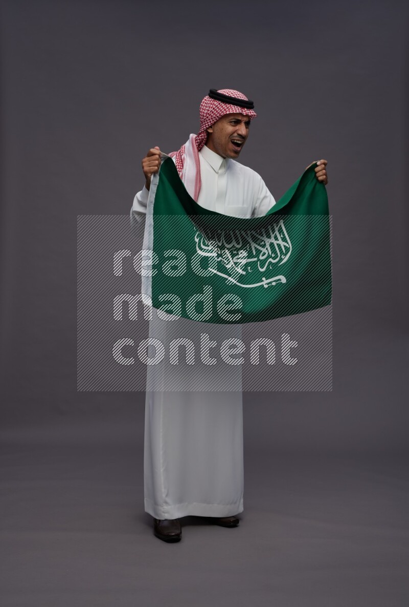 Saudi man wearing thob and shomag standing holding Saudi flag on gray background