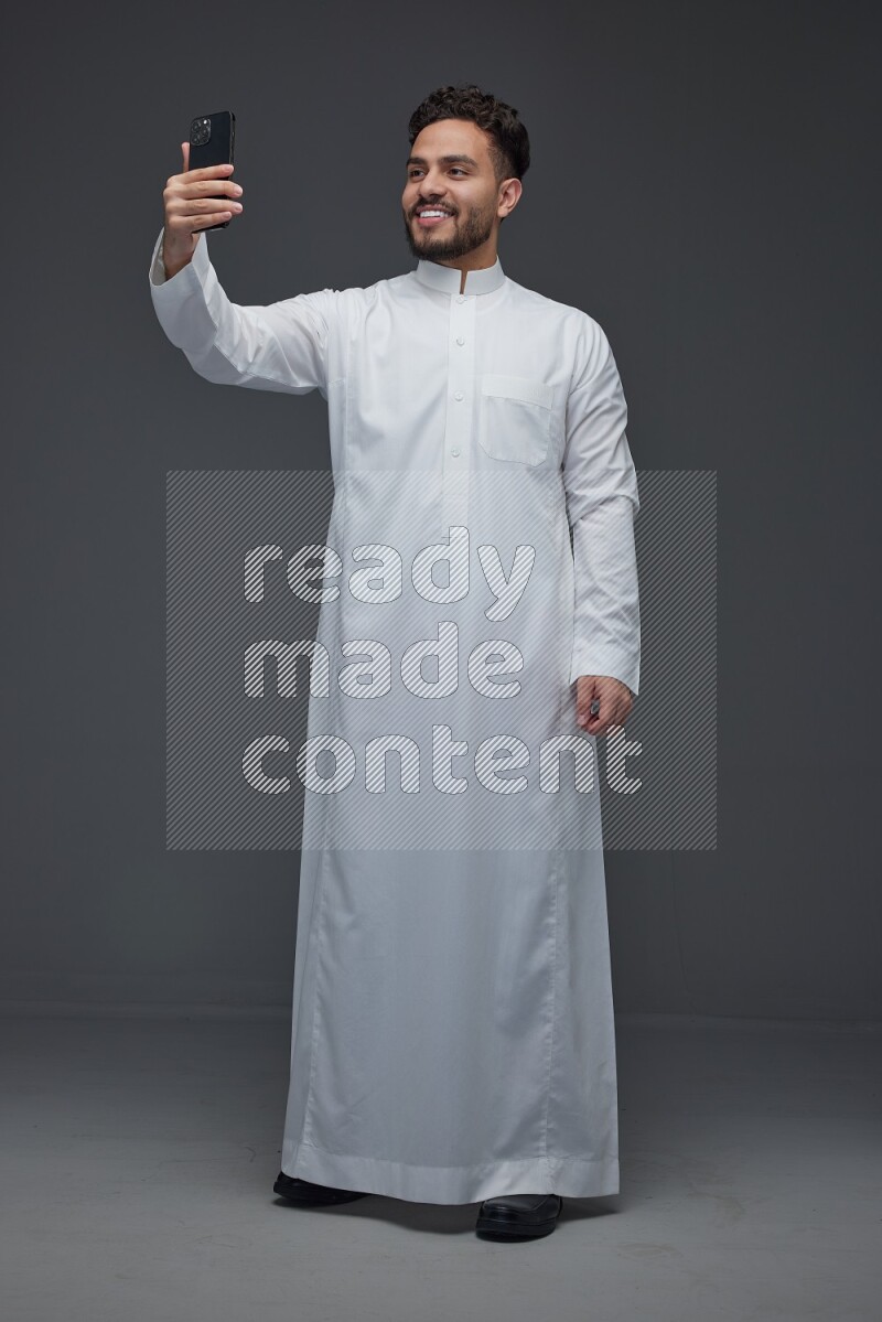 A Saudi man wearing Thobe and taking selfie with his phone making different poses eye level on a gray background