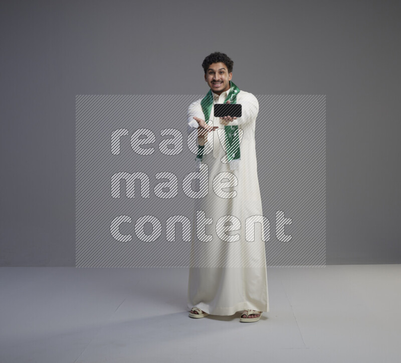 A Saudi man standing wearing thob and Saudi flag scarf showing phone to camera on gray background