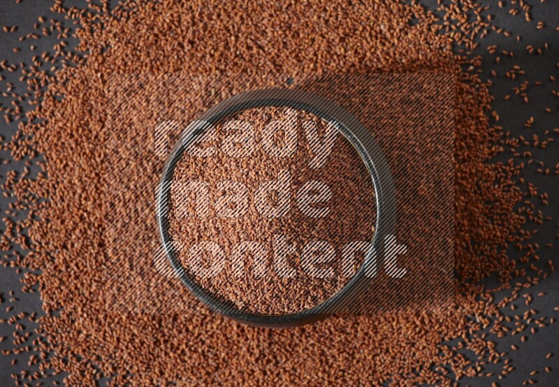 A black pottery bowl full of garden cress seeds surrounded by seeds on a black flooring
