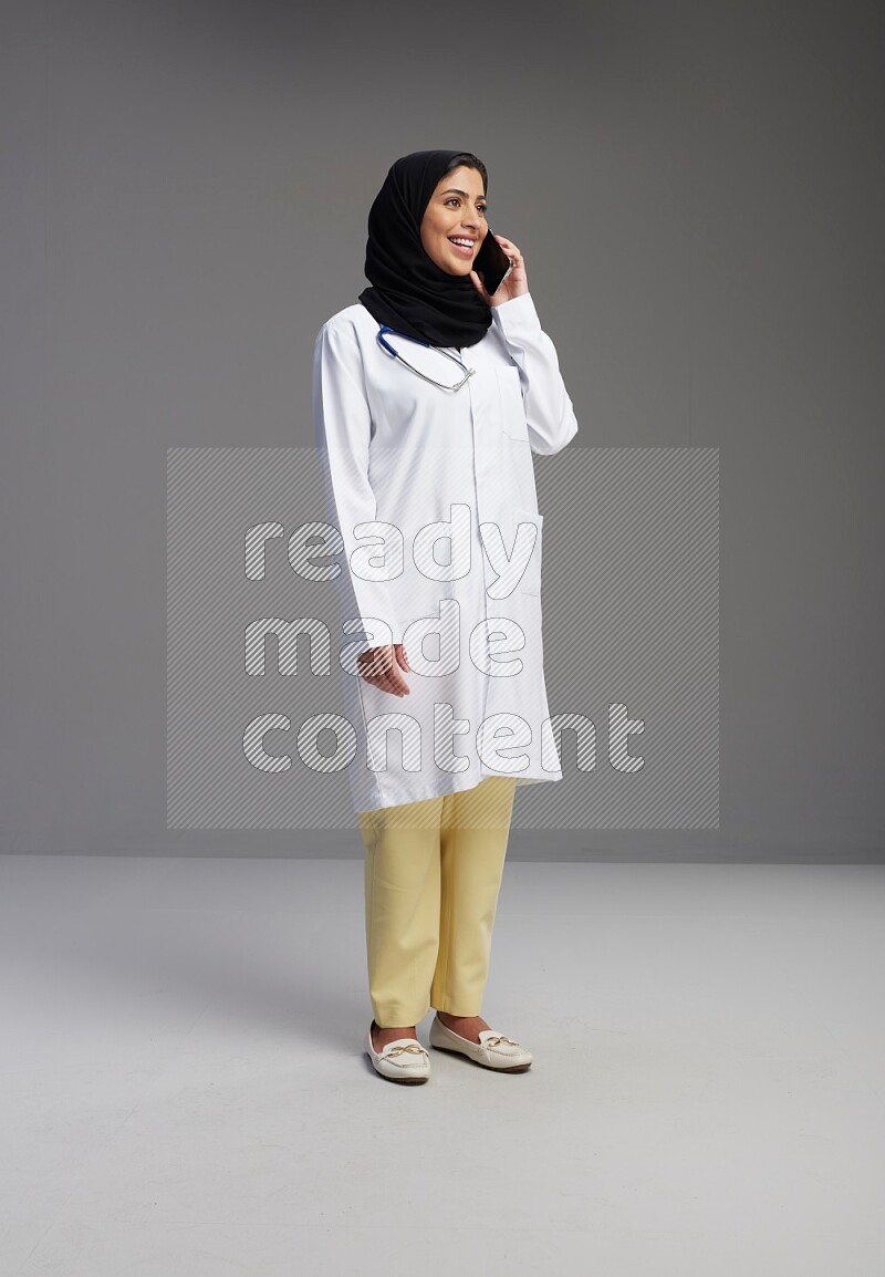 Saudi woman wearing lab coat with stethoscope standing talking on phone on Gray background