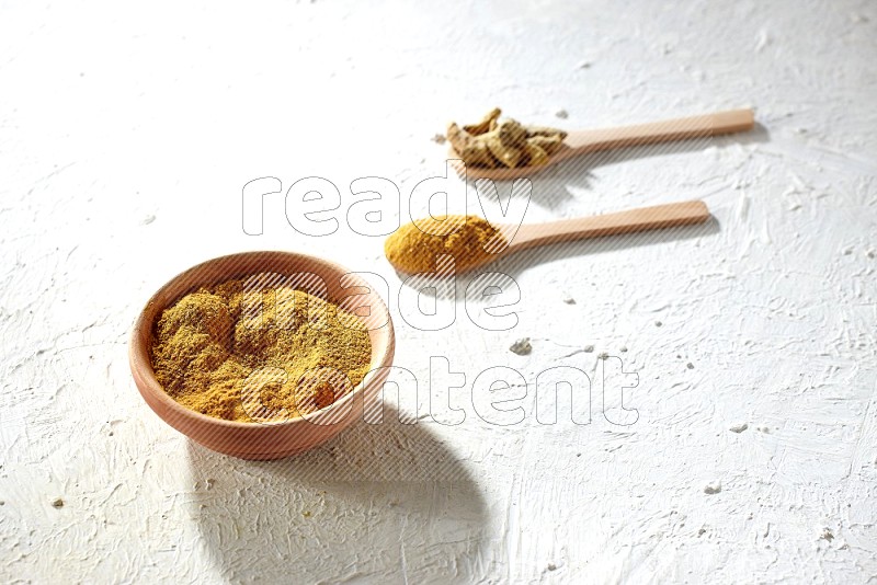 A wooden bowl full of turmeric powder and 2 wooden spoons full of dried turmeric whole finger and turmeric powder on textured white flooring