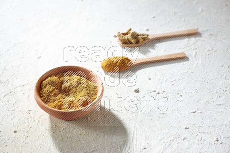 A wooden bowl full of turmeric powder and 2 wooden spoons full of dried turmeric whole finger and turmeric powder on textured white flooring