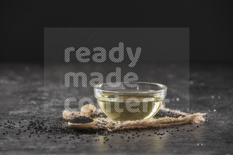 وعاء زجاجي ممتلئ بزيت حبة البركة مع ملعقة خشبية بها بذور حبة البركة مع بعض البذور المتناثرة علي قطعة من القماش علي خلفية سوداء