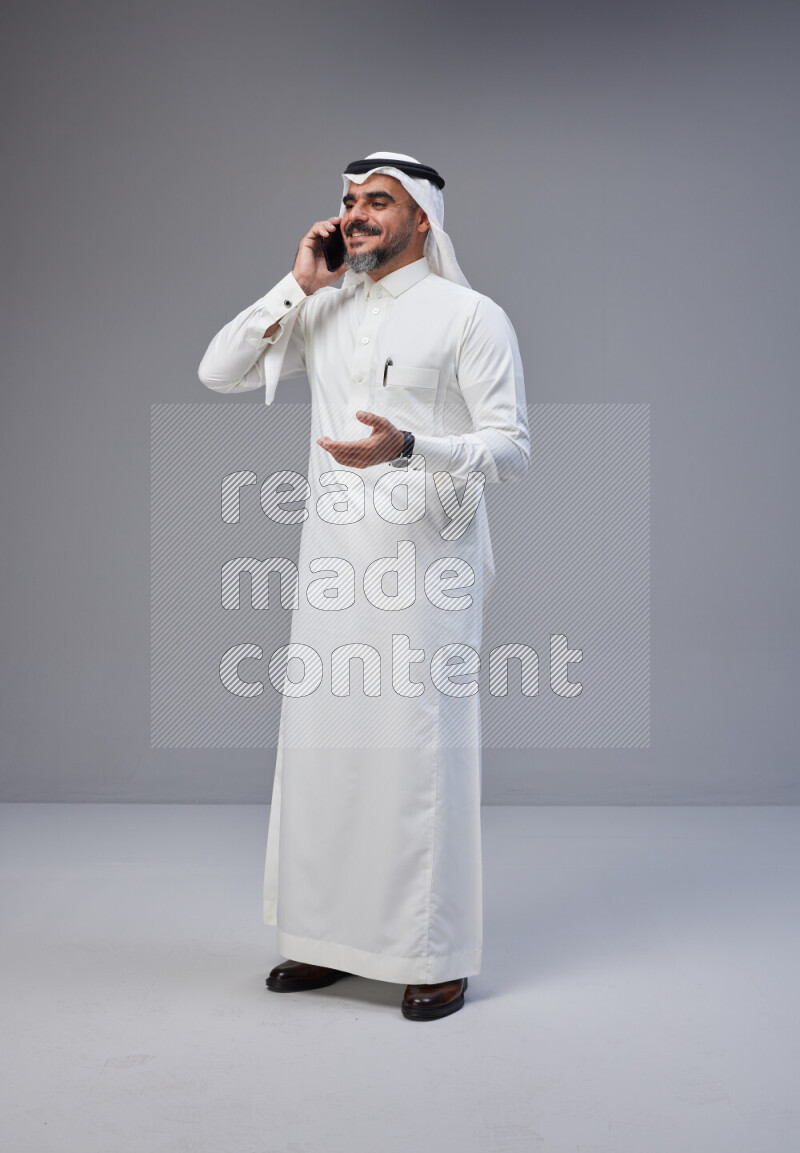 Saudi man Wearing Thob and white Shomag standing talking on phone on Gray background