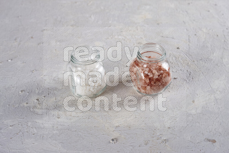 2 glass jars one is filled with coarse himalayan salt and the other with coarse sea salt on white background