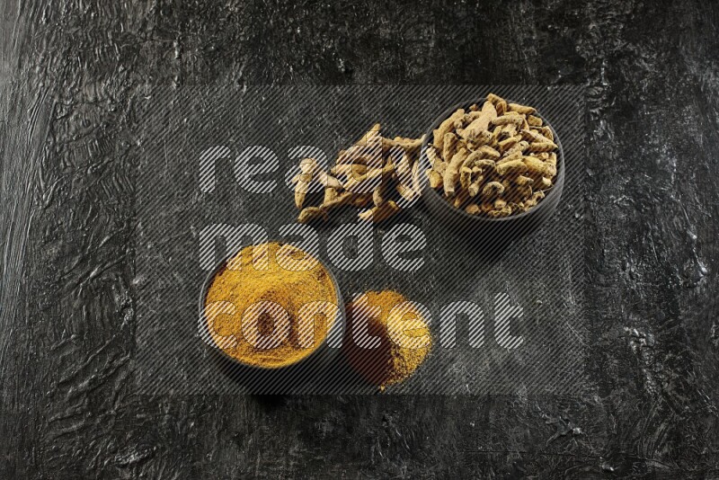 2 pottery black bowls, one full of turmeric powder and the other full of dried turmeric whole fingers on a textured black flooring
