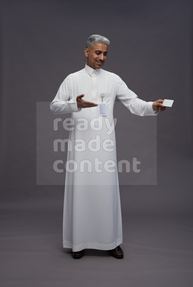 Saudi man wearing thob with pocket employee badge standing holding ATM card on gray background