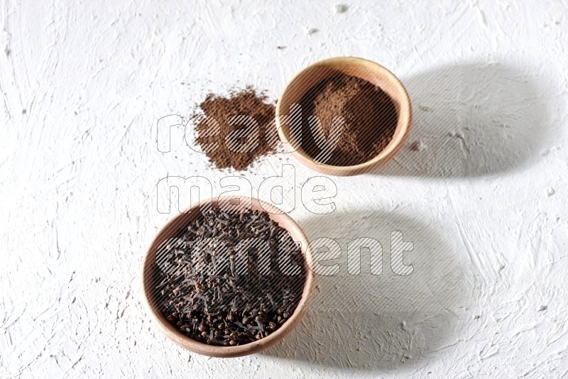 2 wooden bowls full of cloves powder and whole cloves on a textured white flooring