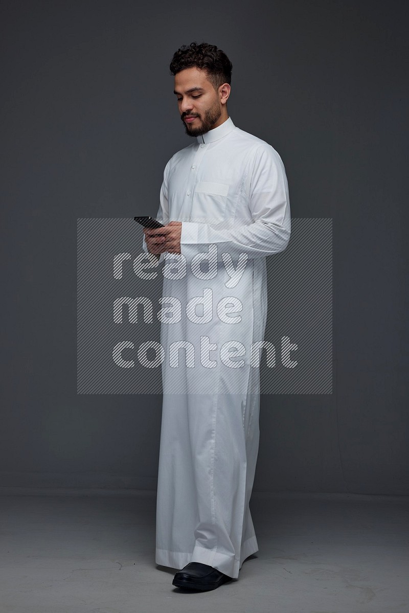 A Saudi man wearing Thobe standing and using his phone eye level on a gray background
