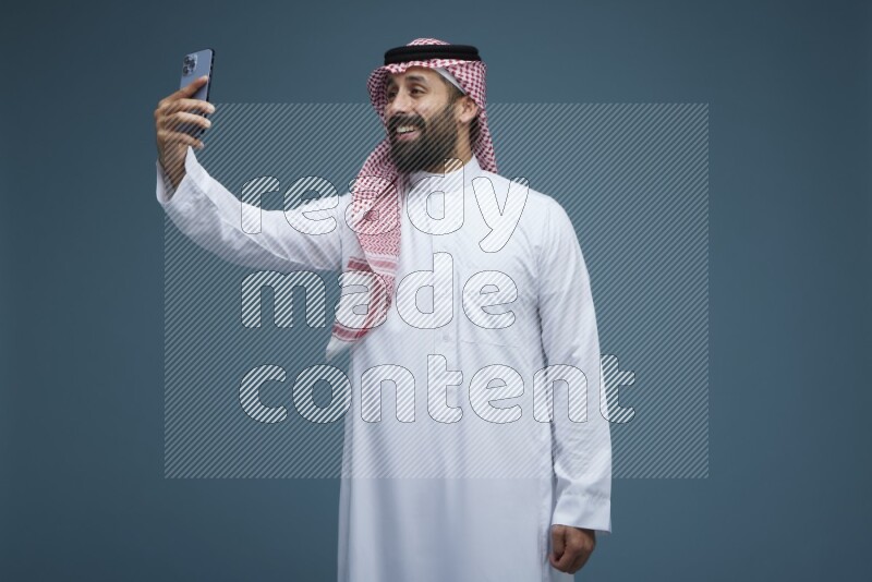 A man Taking Selfie in a blue background wearing Saudi Thob with Shomag