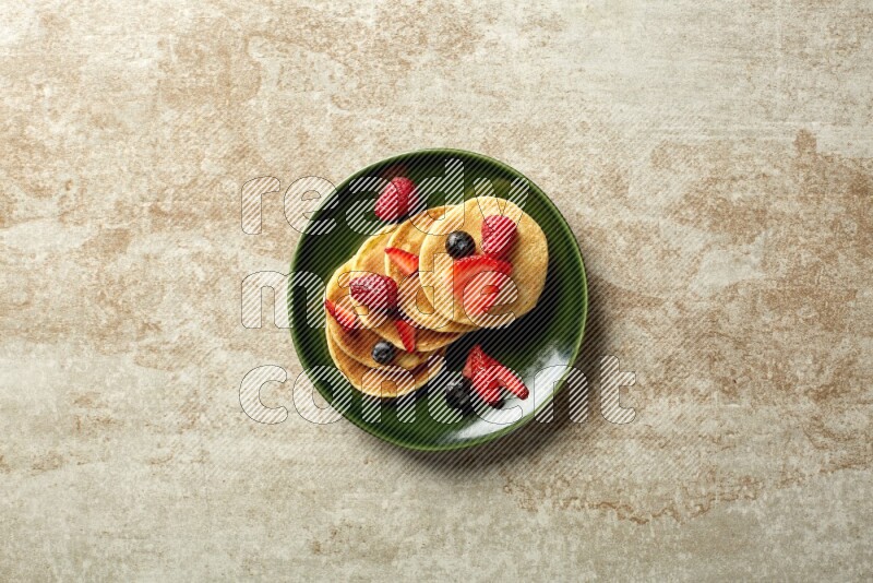 Five stacked mixed berries mini pancakes in a green plate on beige background