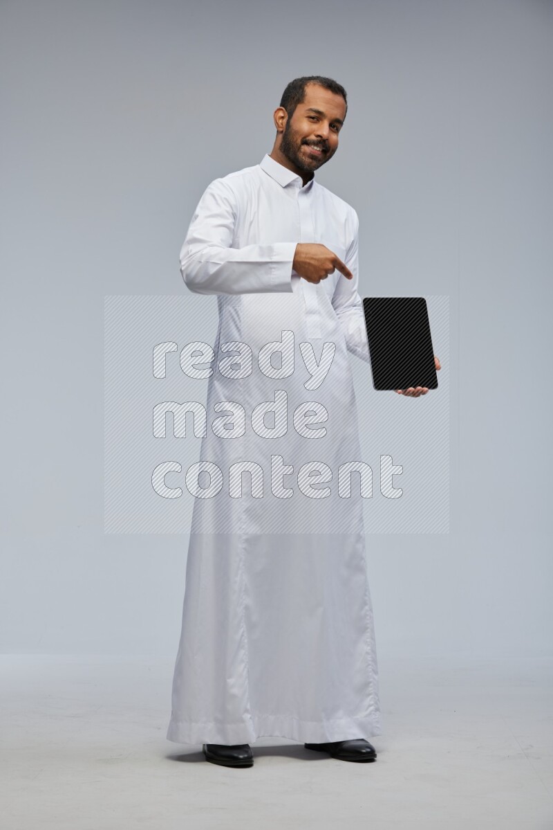 Saudi man wearing Thob standing showing tablet to camera on Gray background