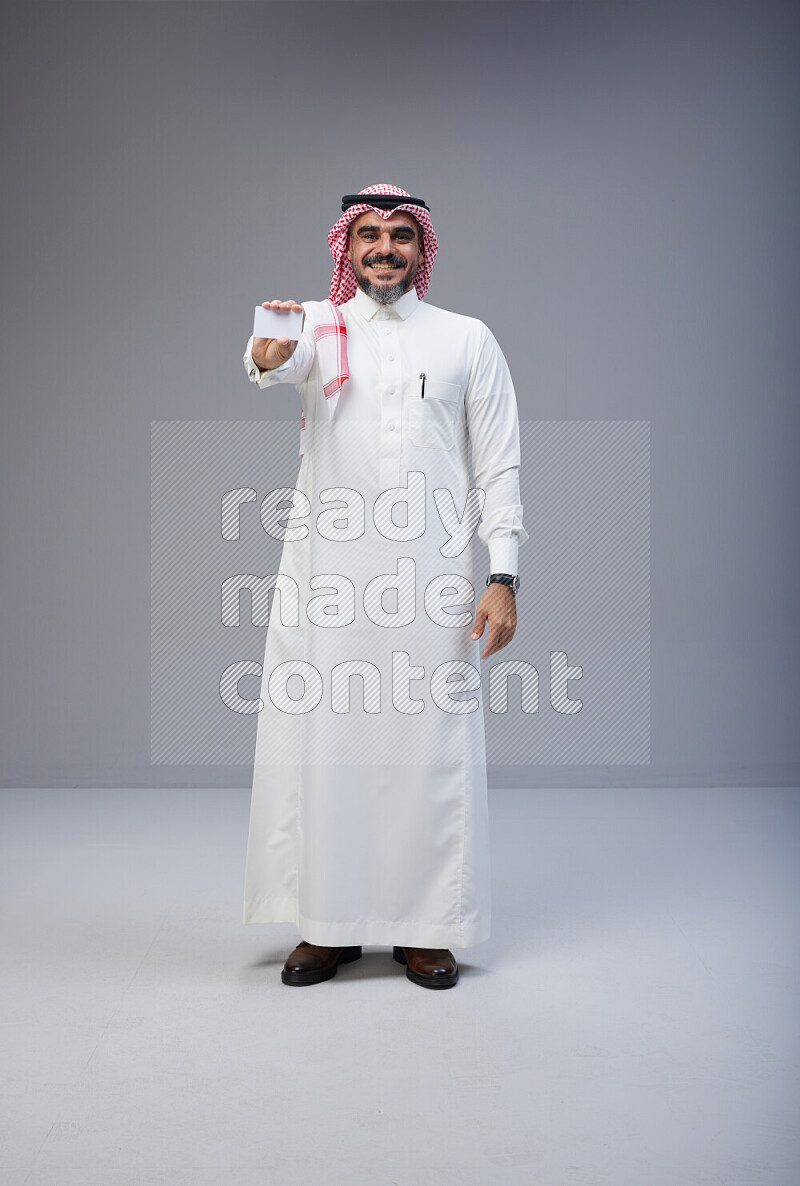 Saudi man Wearing Thob and red Shomag standing holding ATM card on Gray background