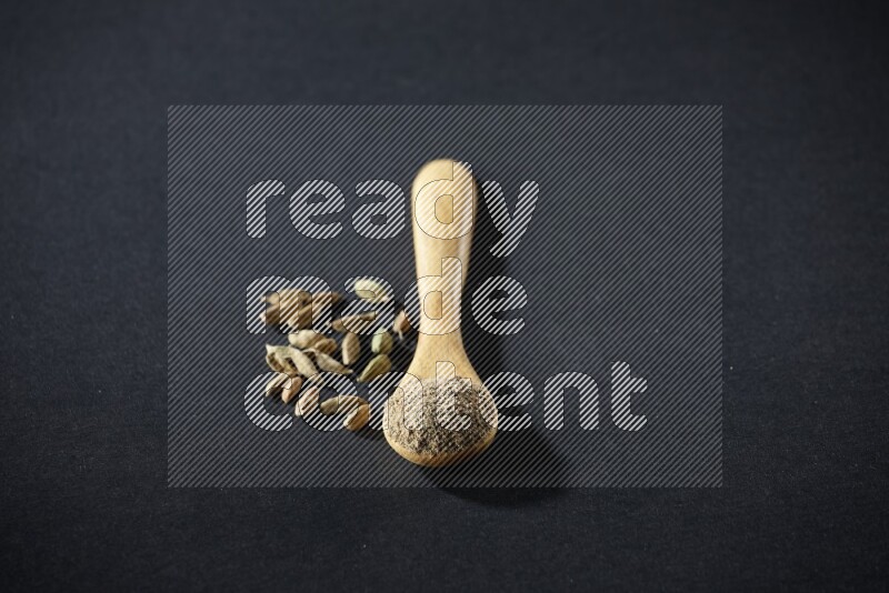A wooden spoon full of cardamom powder and cardamom seeds beside it on black flooring