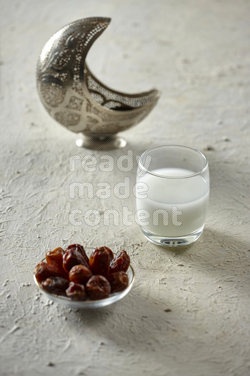 A silver lantern with different drinks, dates, nuts, prayer beads and quran on textured white background