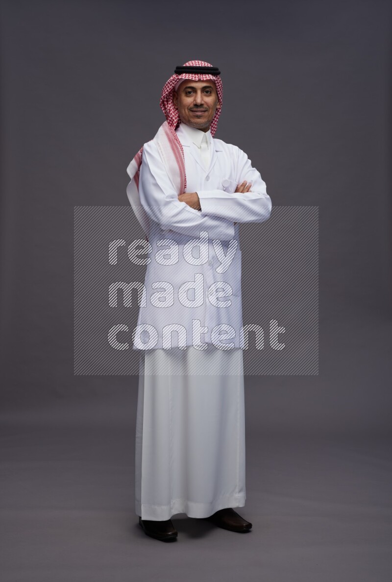 Saudi man wearing thob with lab coat and shomag with pocket employee badge standing with crossed arms on gray background
