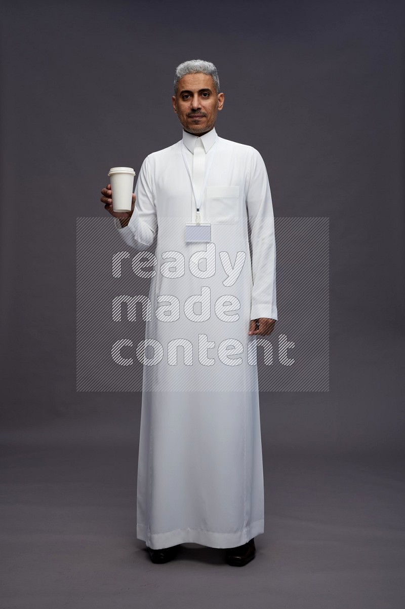 Saudi man wearing thob with neck strap employee badge standing holding paper cup on gray background