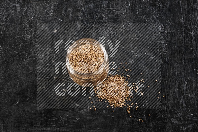 A glass jar full of mustard seeds and jar is flipped and seeds spread out on a textured black flooring in different angles