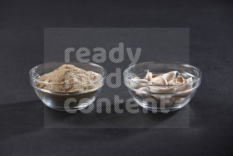 2 glass bowls, one full of garlic powder and the other full of garlic cloves on a black flooring