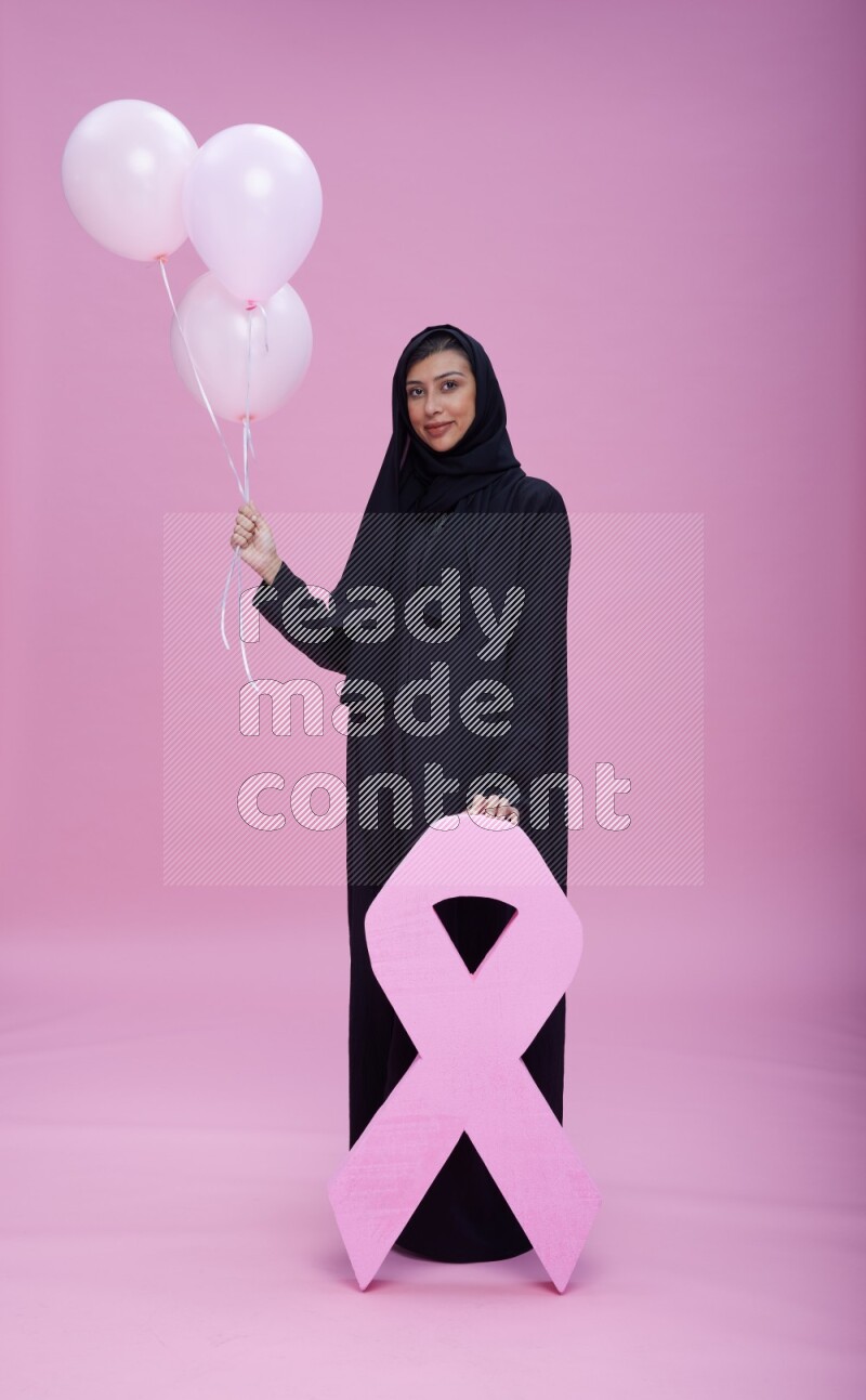 Saudi woman wearing pink ribbon on Abaya standing holding balloons and awareness sign on pink background