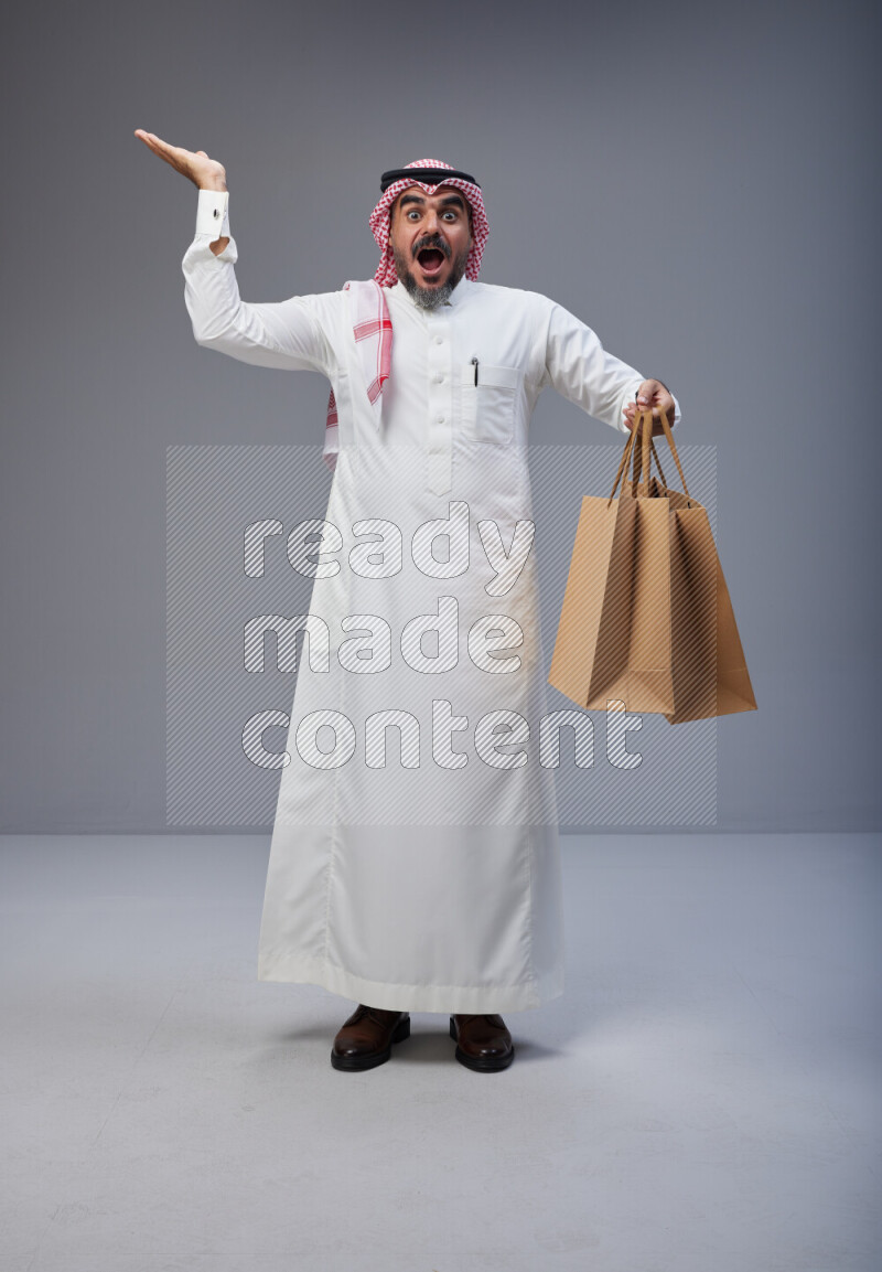 Saudi man Wearing Thob and red Shomag standing holding shopping bag on Gray background