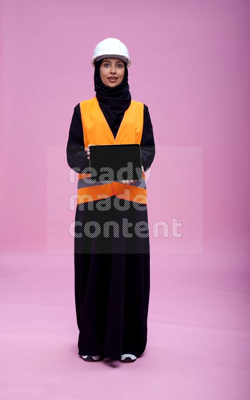 Saudi woman wearing Abaya with engineer vest and helmet standing showing tablet to camera on pink background