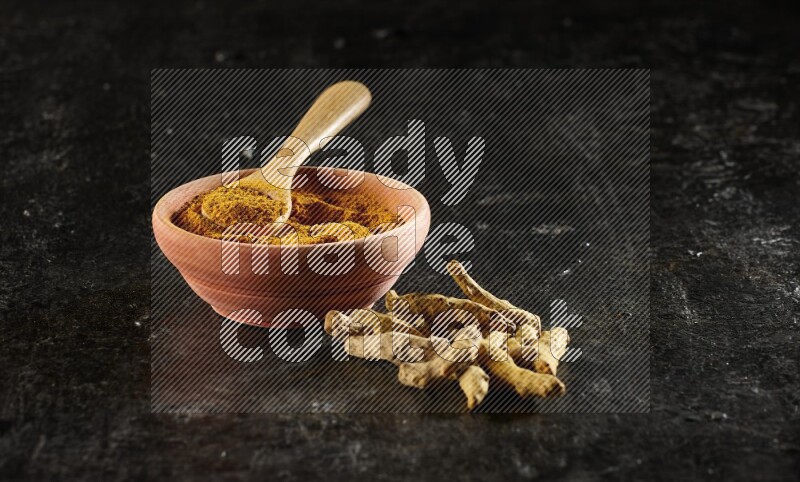 A wooden bowl and a wooden spoon full of turmeric powder with dried turmeric fingers on textured black flooring