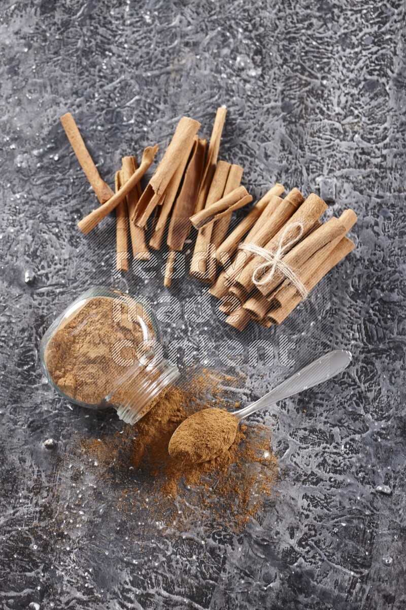 Herbal glass jar full cinnamon powder flipped and a metal spoon full of powder, cinnamon sticks stacked and bounded in the back on textured black background in different angles