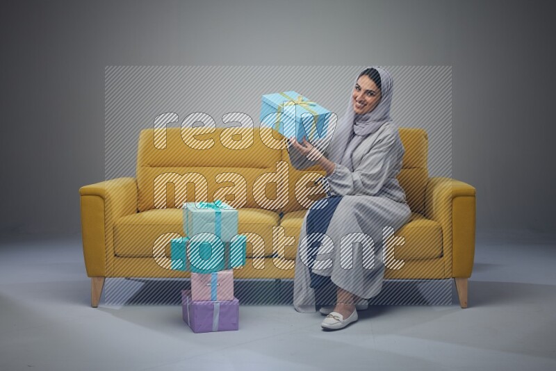 A Saudi woman wearing a light gray Abaya and head scarf sitting on a yellow sofa holding a gift box beside multi color gift boxes eye level on a grey background