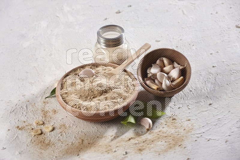 A wooden bowl, spoon and glass spice jar full of garlic powder and a wooden bowl full of garlic cloves on a textured white flooring in different angles