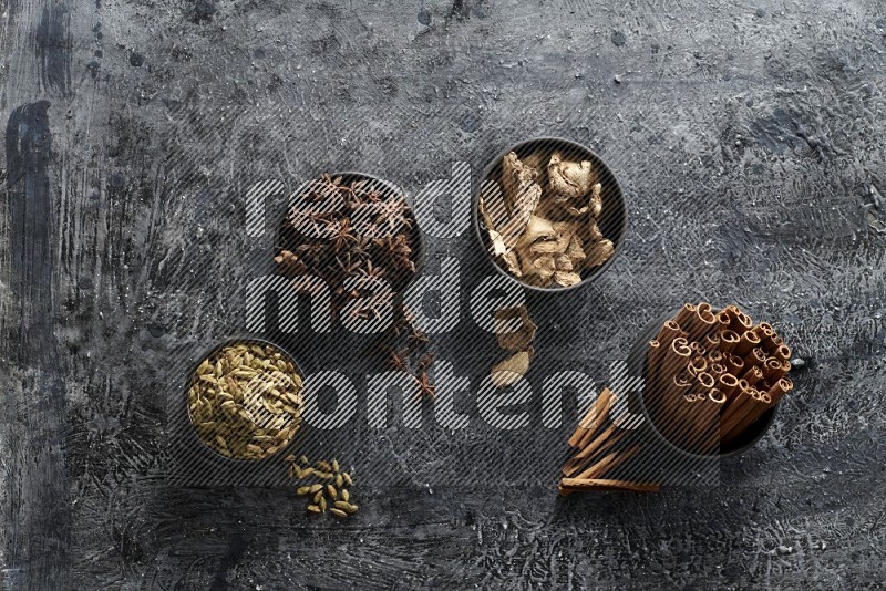 Ginger, Cardamom, Star anise and cinnamon sticks in 4 bowls on a textured black background