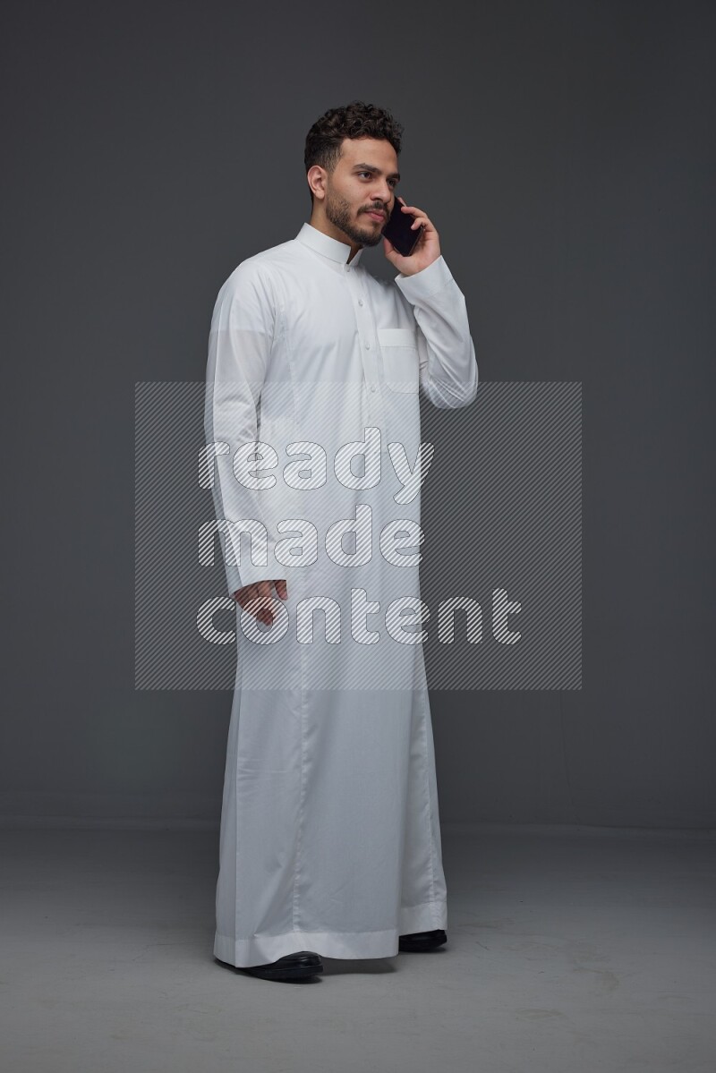 A Saudi man wearing Thobe and talking in the phone while standing and making different poses eye level on a gray background
