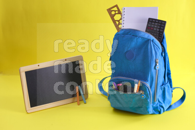 A school bag with assorted school supplies in and beside it on yellow background