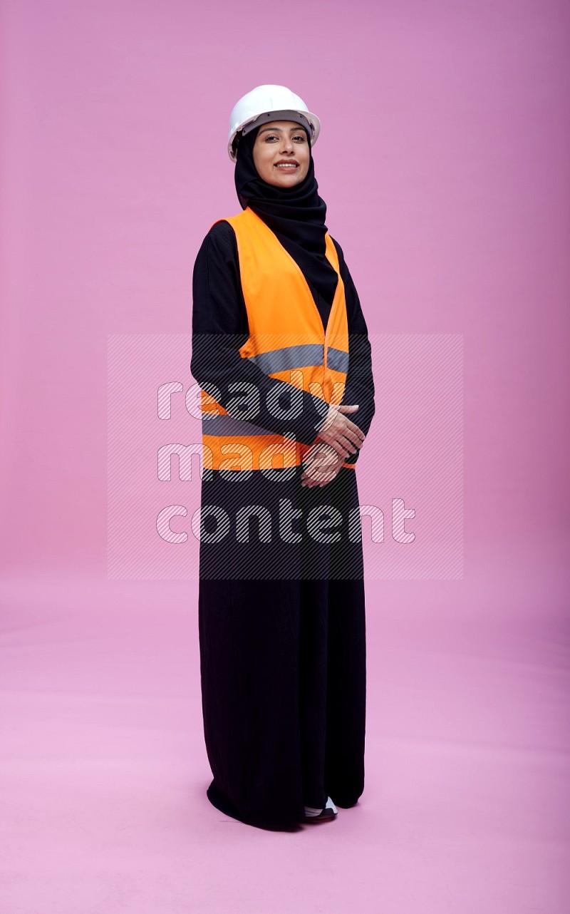 Saudi woman wearing Abaya with engineer vest and helmet standing interacting with the camera on pink background