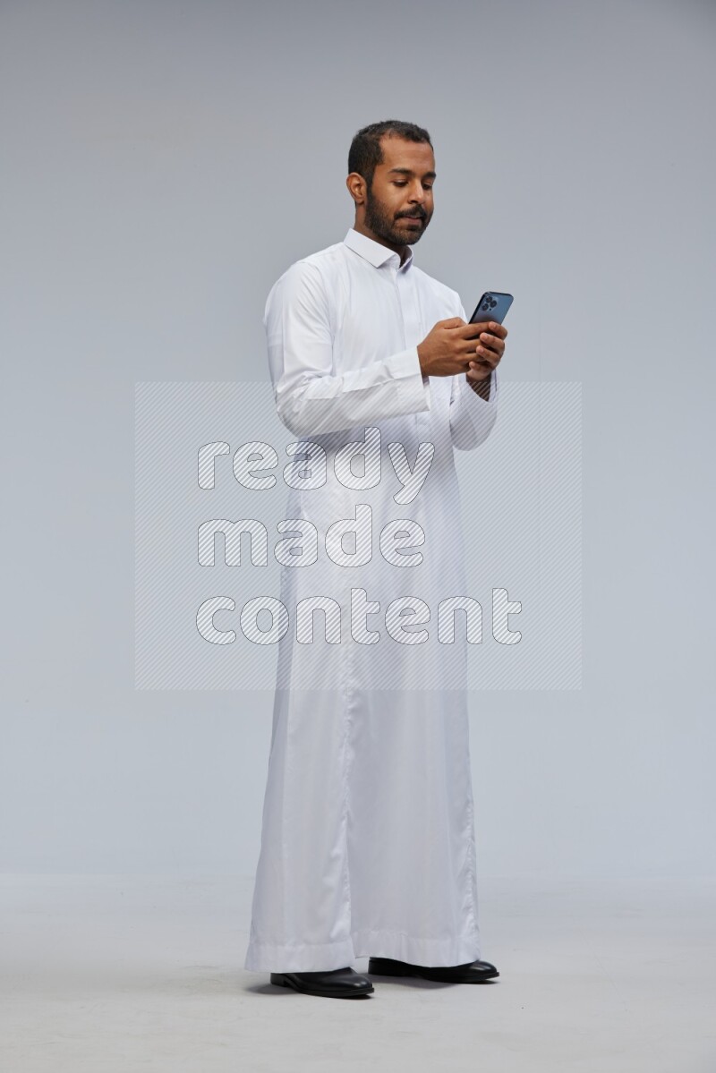 Saudi man wearing Thob standing texting on phone on Gray background