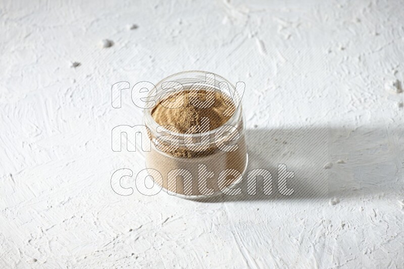 A glass jar full of cumin powder on textured white flooring