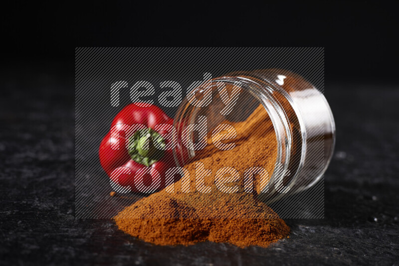 A glass jar full of ground paprika powder flipped with some spilling powder on black background