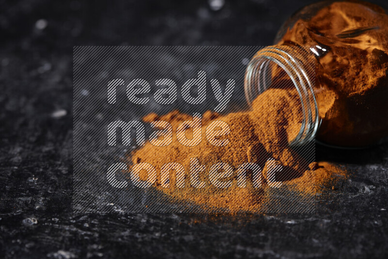 A glass jar full of ground paprika powder flipped with some spilling powder on black background