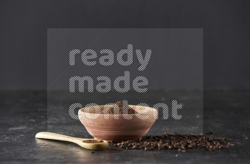 A wooden bowl and wooden spoon full of cloves powder with spreaded cloves on textured black flooring