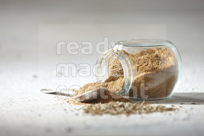 A glass spice jar and metal spoon full of cumin powder and the jar flipped and powder spilled out with cumin seeds on textured white flooring