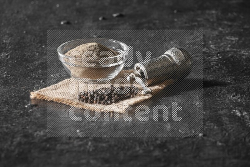 A glass bowl full of black pepper powder and black pepper beads on burlap fabric with a turkish metal pepper grinder on textured black flooring