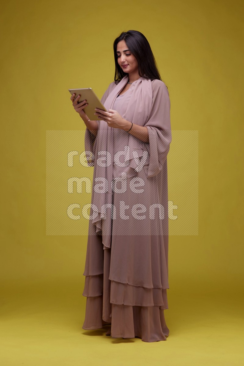 A Saudi woman Holding an iPad  on a Yellow Background wearing Brown Abaya